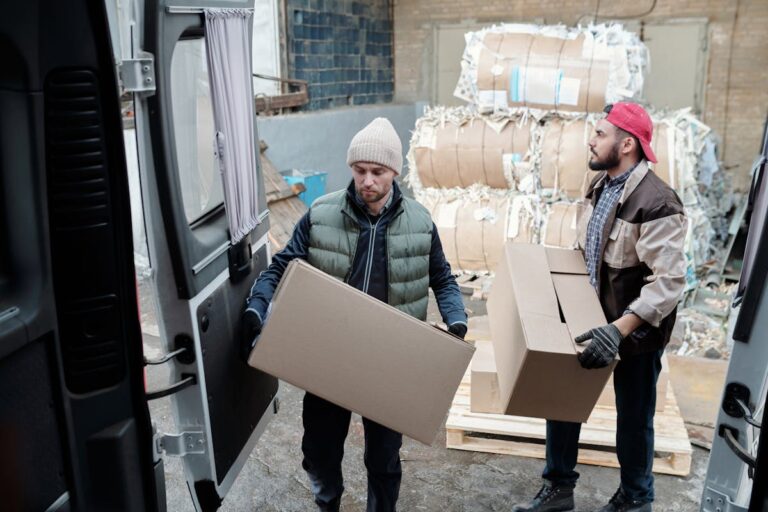 Two male workers loading cardboard boxes in a warehouse with bundled paper in the background