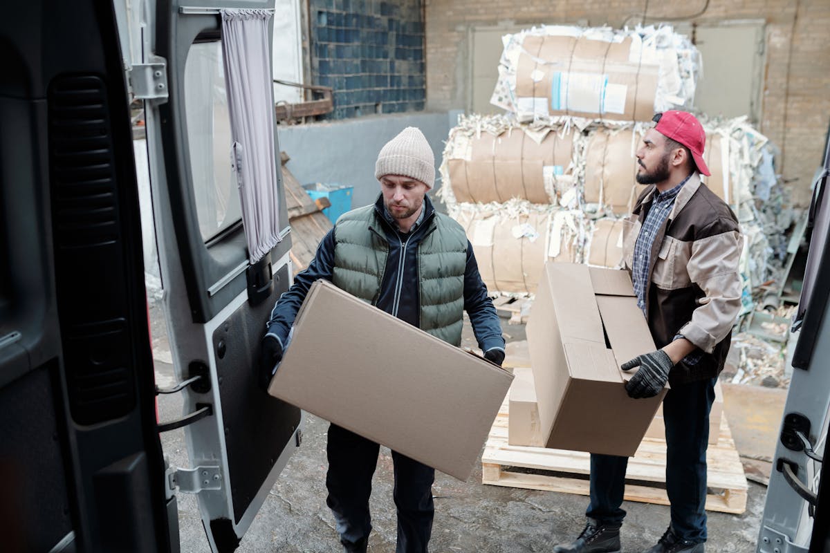 Two male workers loading cardboard boxes in a warehouse with bundled paper in the background